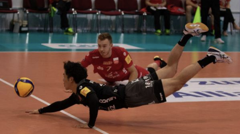 Zwei Volleyballspieler stürzen auf dem Feld, einer im schwarzen Trikot erreicht den Ball.